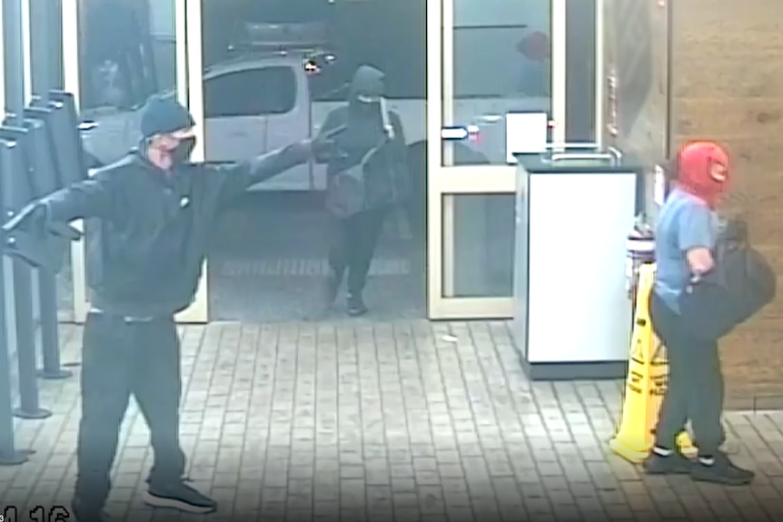 man in black with a black mask holds what appears to be a gun at a fast-food cash register, two women with masks enter store