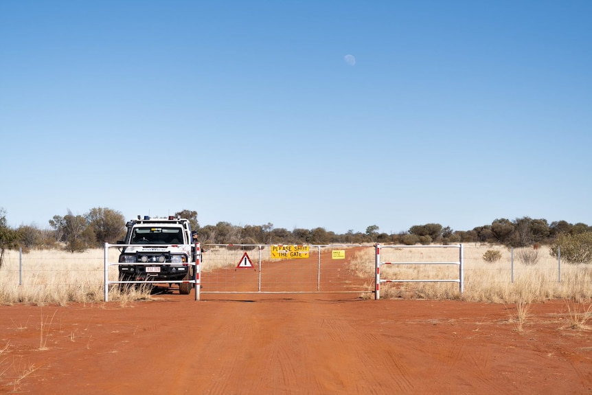 A red dirt road with a cattle gate across it and a parked police truck on the other side.