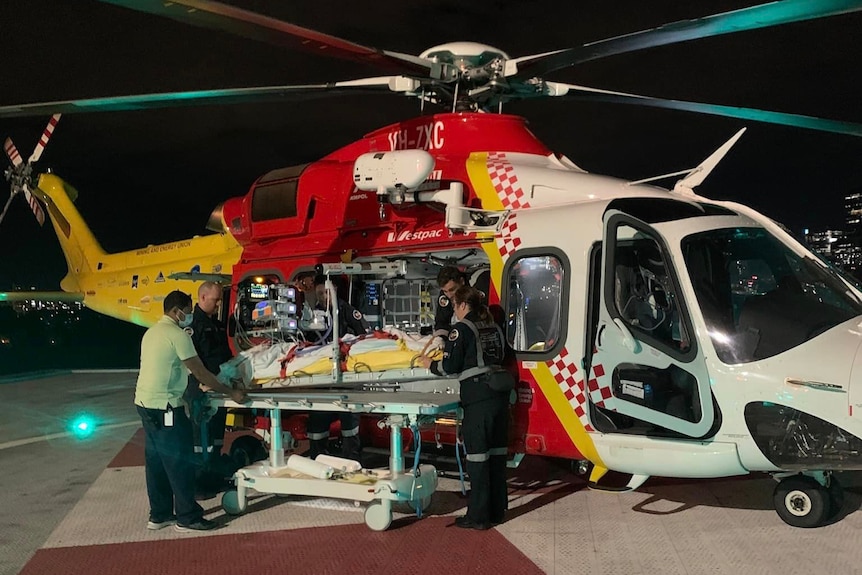 A patient on a bed surrounded by paramedics in front of a rescue helicopter.