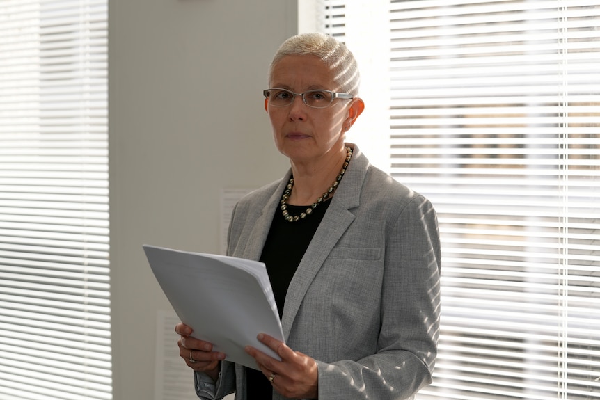 A woman wears a grey jacket holding a stack of files near a window with blinds down.