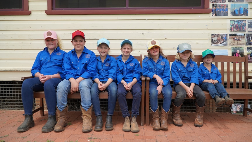 Seven primary school kids wearing work gear sit on a park bench in front of a timber school building.