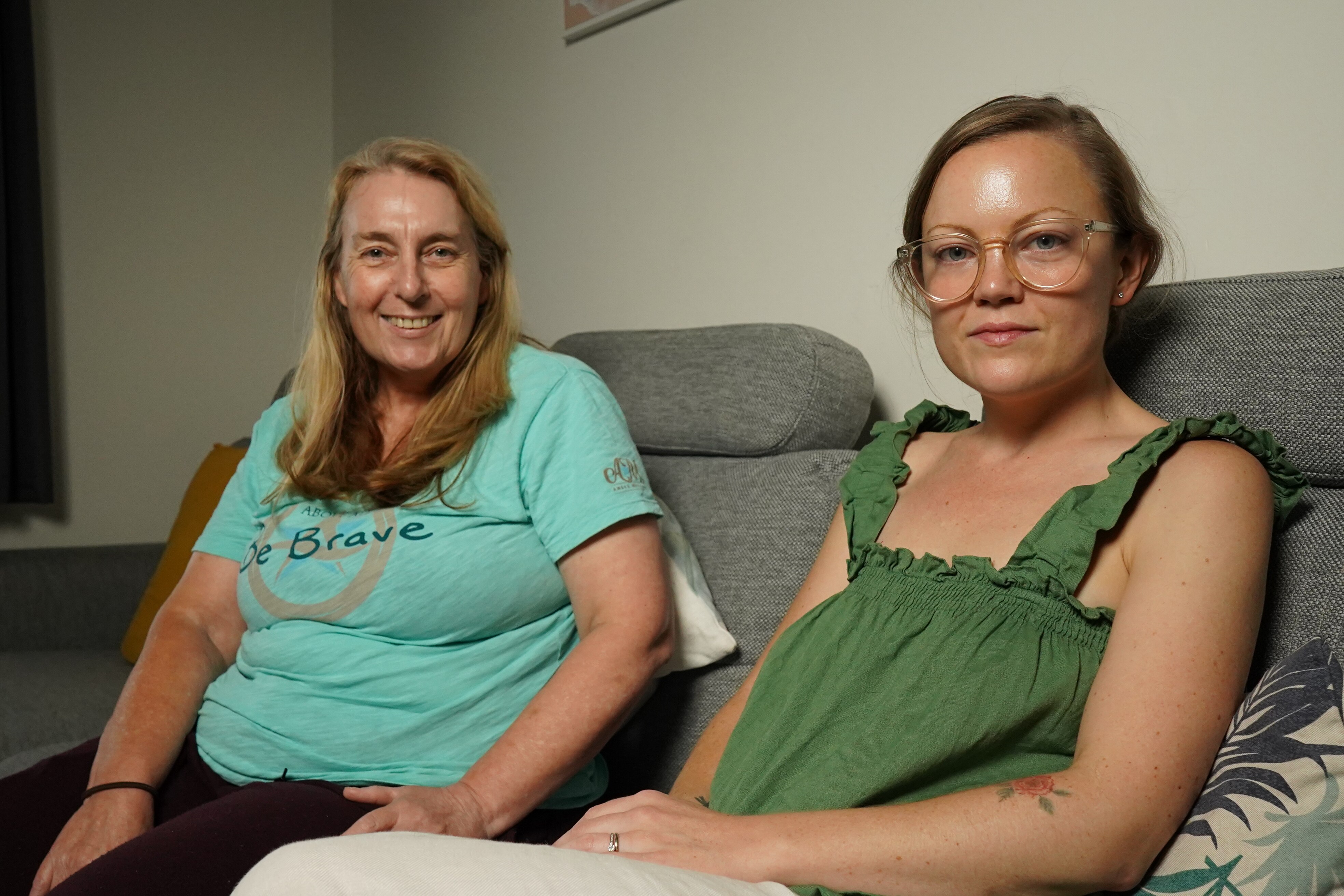 An older woman in a light green t-shirt sits on a couch next to a younger woman in a green top.