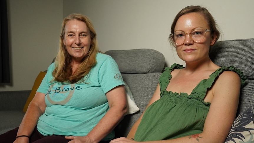 An older woman in a light green t-shirt sits on a couch next to a younger woman in a green top.