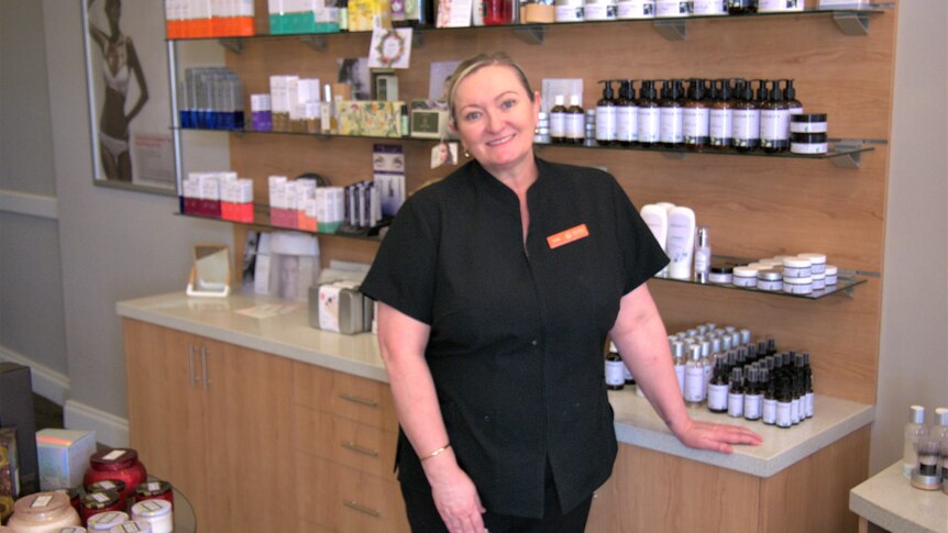 A woman in a black uniform stands smiling in front of shelves of beauty products.