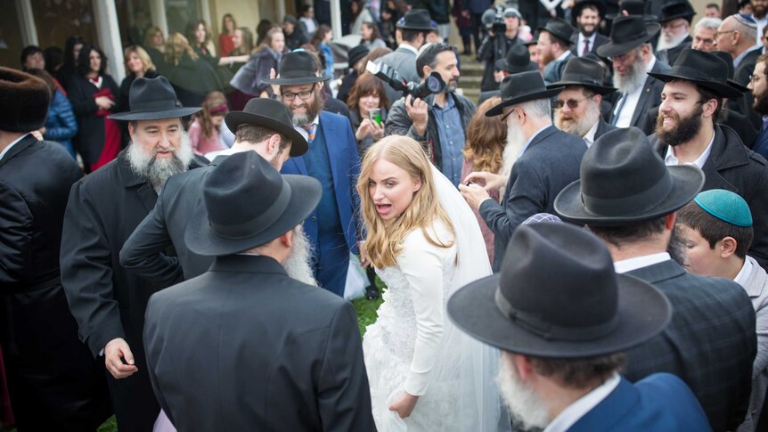 A bride in a wedding dress looks back, surrounded by a crowd of bearded Jewish men in hats.