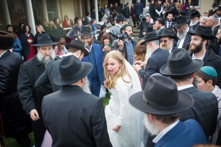 A bride in a wedding dress looks back, surrounded by a crowd of bearded Jewish men in hats.