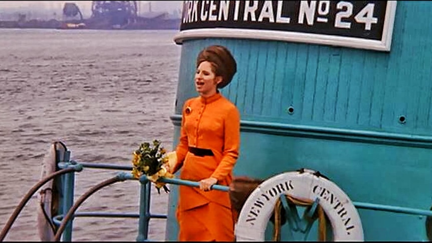 Barbra Streisand wears an orange tailored dress, as she sings at the front of a ferry travelling on the water. 