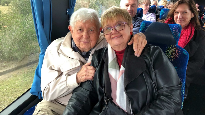An elderly man and his wife sitting on a coach with other passengers in the background