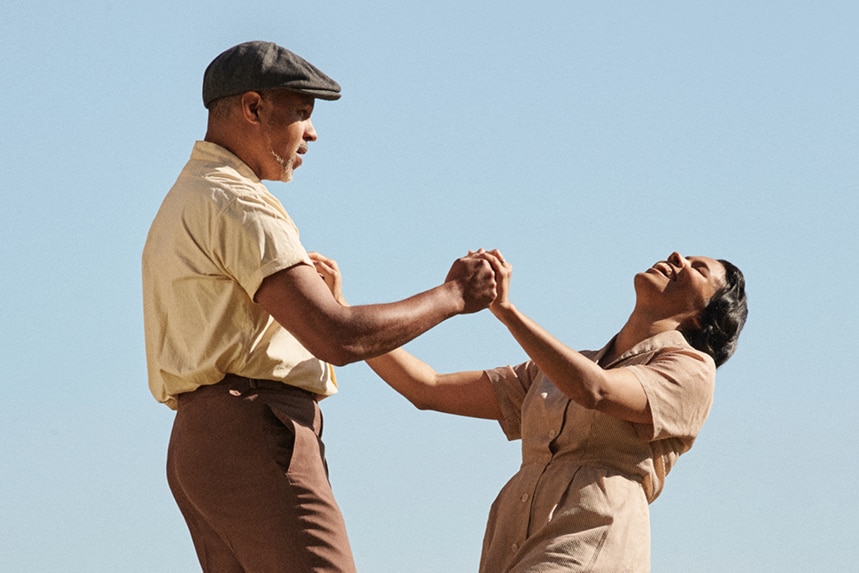 A Black man and woman hold hands as they dance. They each wear 50s-style clothing, the woman is leaned back with a wide smile.