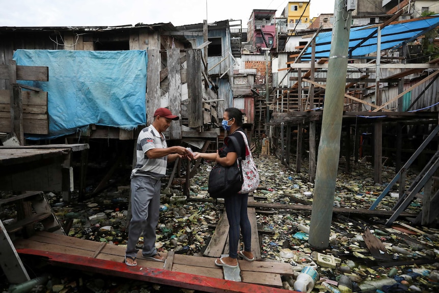 A man hands over a mask to a woman on a piece of wood against a background of run-down housing