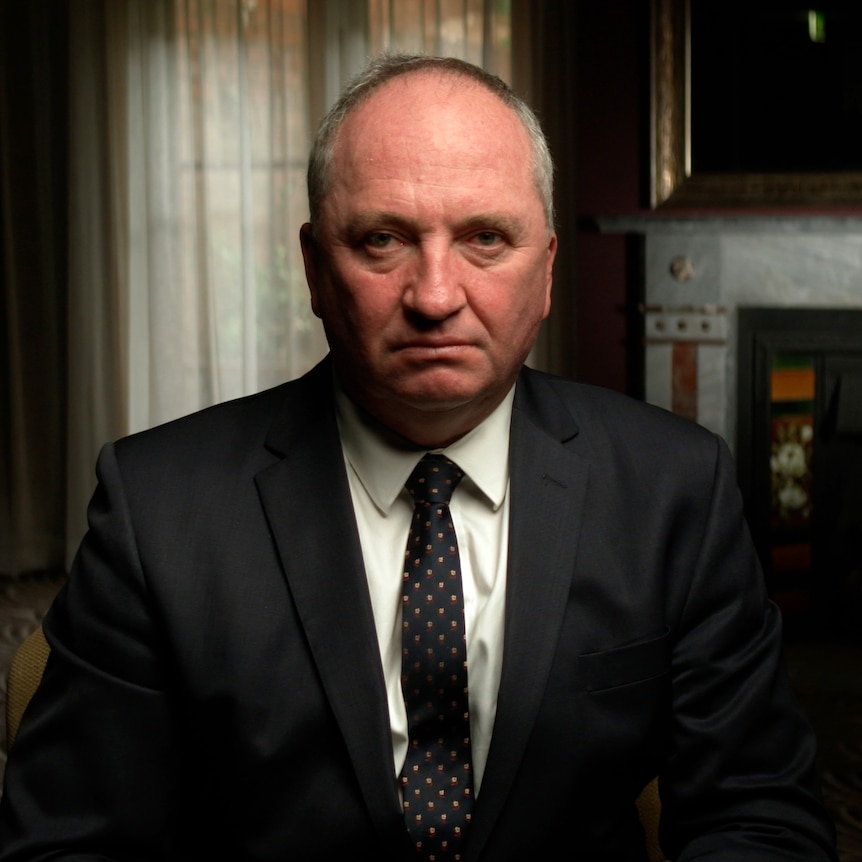 Dressed in a dark suit and tie, Barnaby Joyce sits in a room, looking into camera with a serious expression.