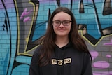 A teenage girl with long brown hair standing in front of a wall decorated in graffiti.