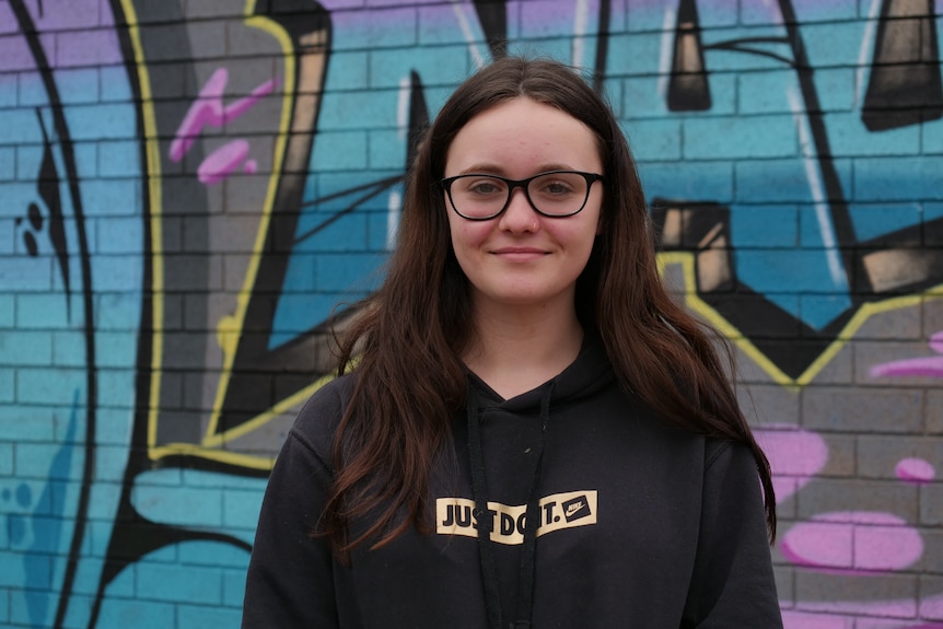 A teenage girl with long brown hair standing in front of a wall decorated in graffiti.