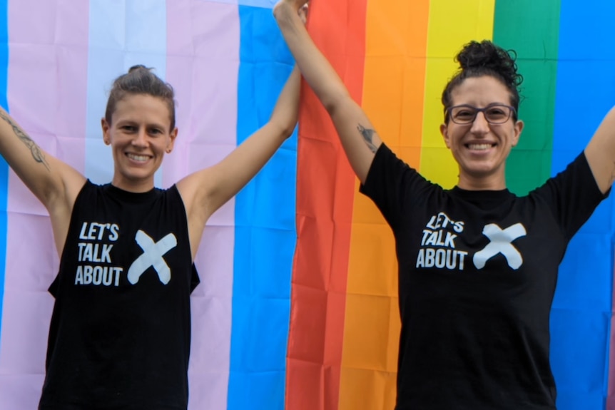 Two people wearing black shirts that say Let's Talk About X stand in front of pride flags. They are smiling and look proud.