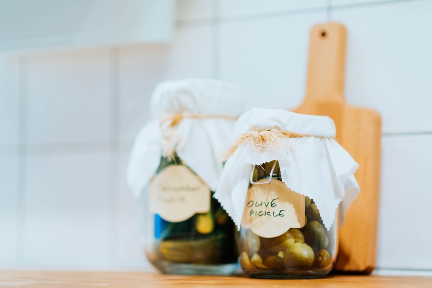 Two labelled jars of olive pickles in front of a chopping board on a kitchen bench for a story about pickling.