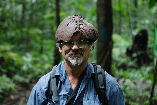 A man with a backpack and glasses wears a half rotten old fruit shell on his head in the rainforest.
