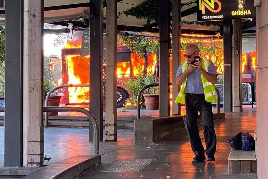 a bus driver is on the phone and a bus is completely engulfed in flames in the background