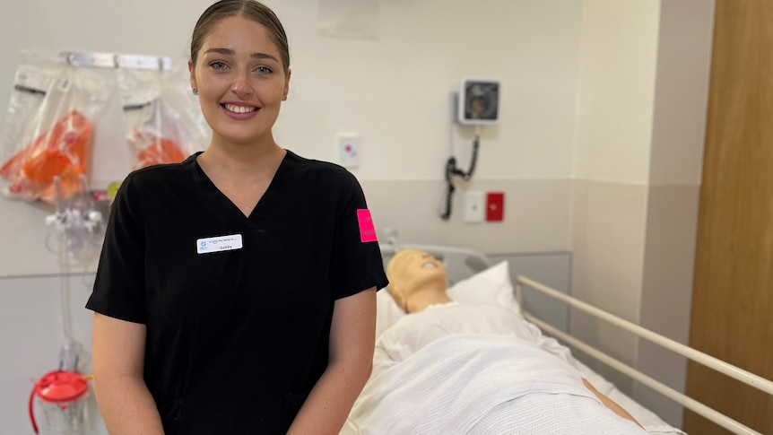 Young woman in nurse uniform smiles, standing in front of hospital bed with dummy patient