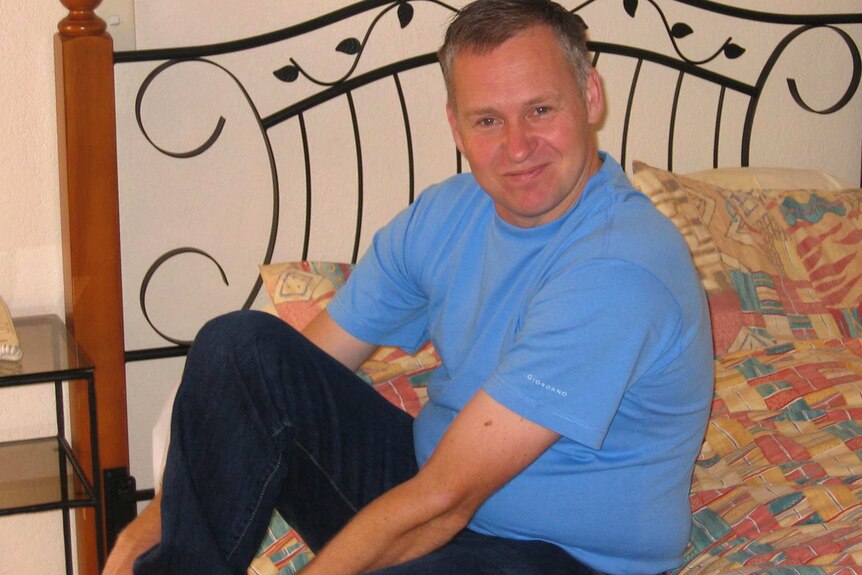 A man wearing a blue shirt sits smiling on a bed while putting a shoe on.
