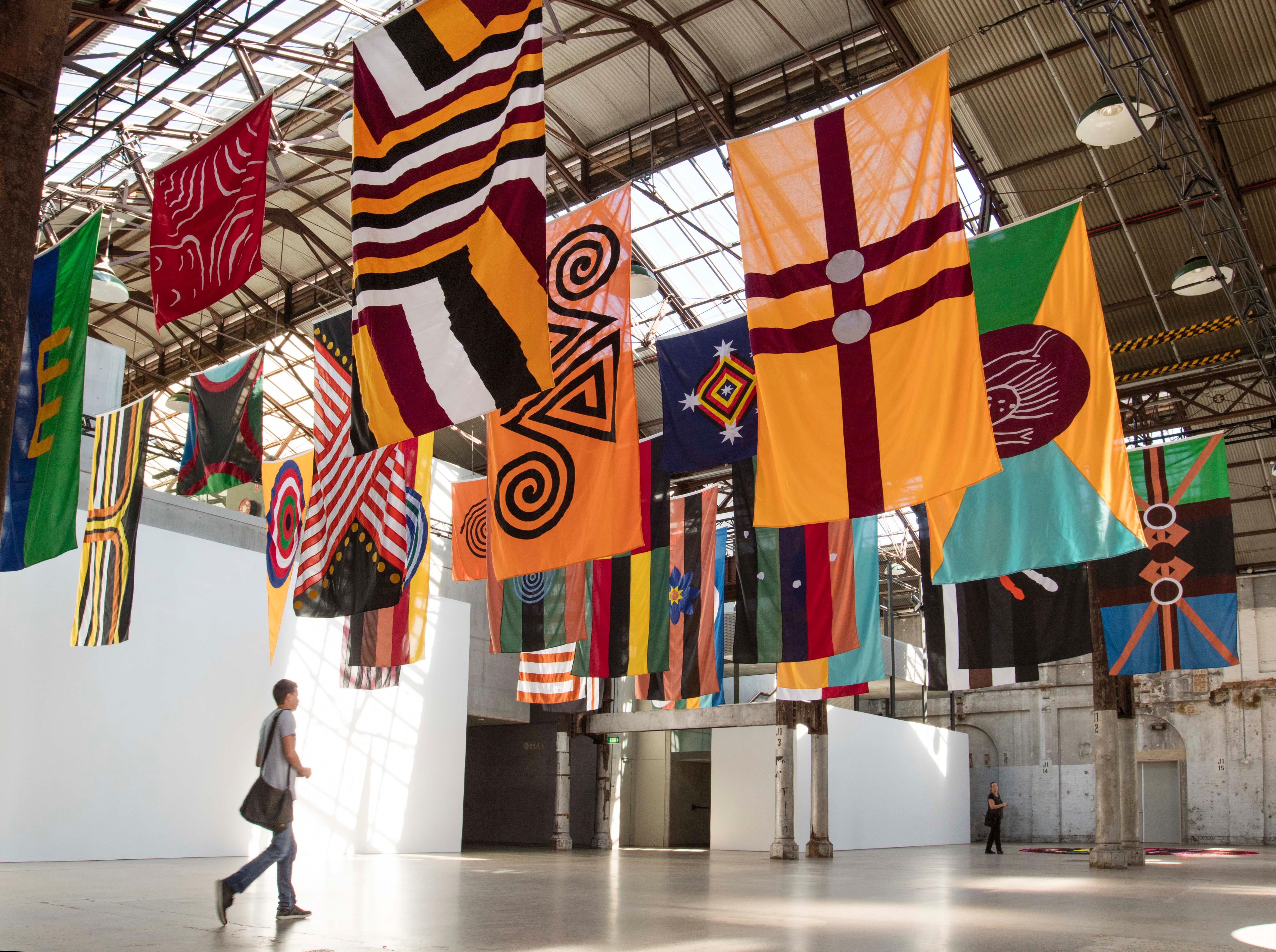 A series of colourful flags depicting First Nations designs and symbols hang from the ceiling of a industrial gallery space.