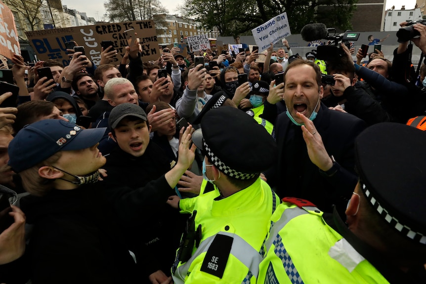 A crowd of football fans shout as Petr Cech wears a face mask and pleads for peace