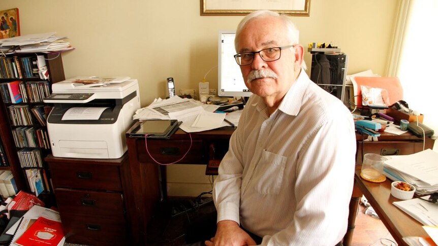 Dr Stephen Duckett sits at a desk cluttered with papers, books and stationary and a computer.