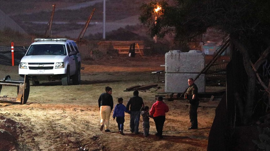 Three adults and two children walk in a line holding hands, as a uniformed man looks on, at night, near a large vehicle