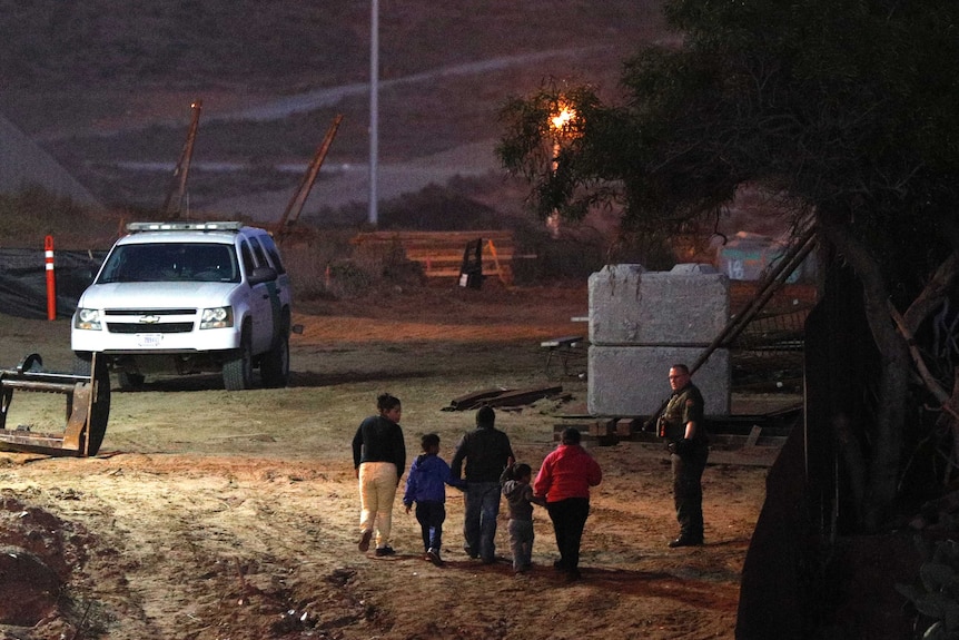 Three adults and two children walk in a line holding hands, as a uniformed man looks on, at night, near a large vehicle