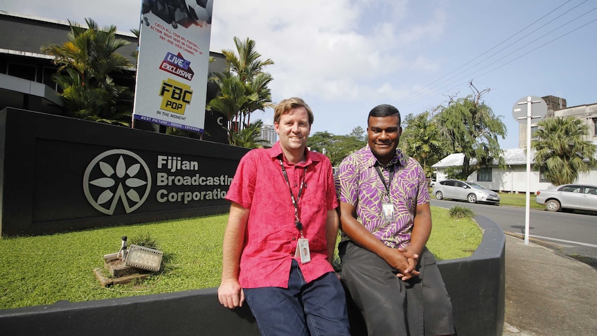 Two men sitting outside smiling in front of the Fijian Broadcasting Corporation