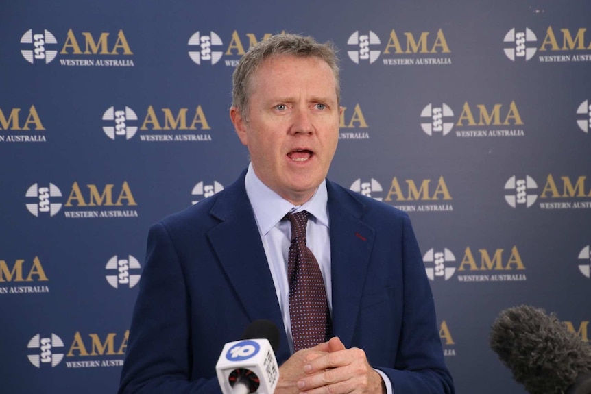 A man wearing a blue suit and tie is speaking in front of an AMA WA banner