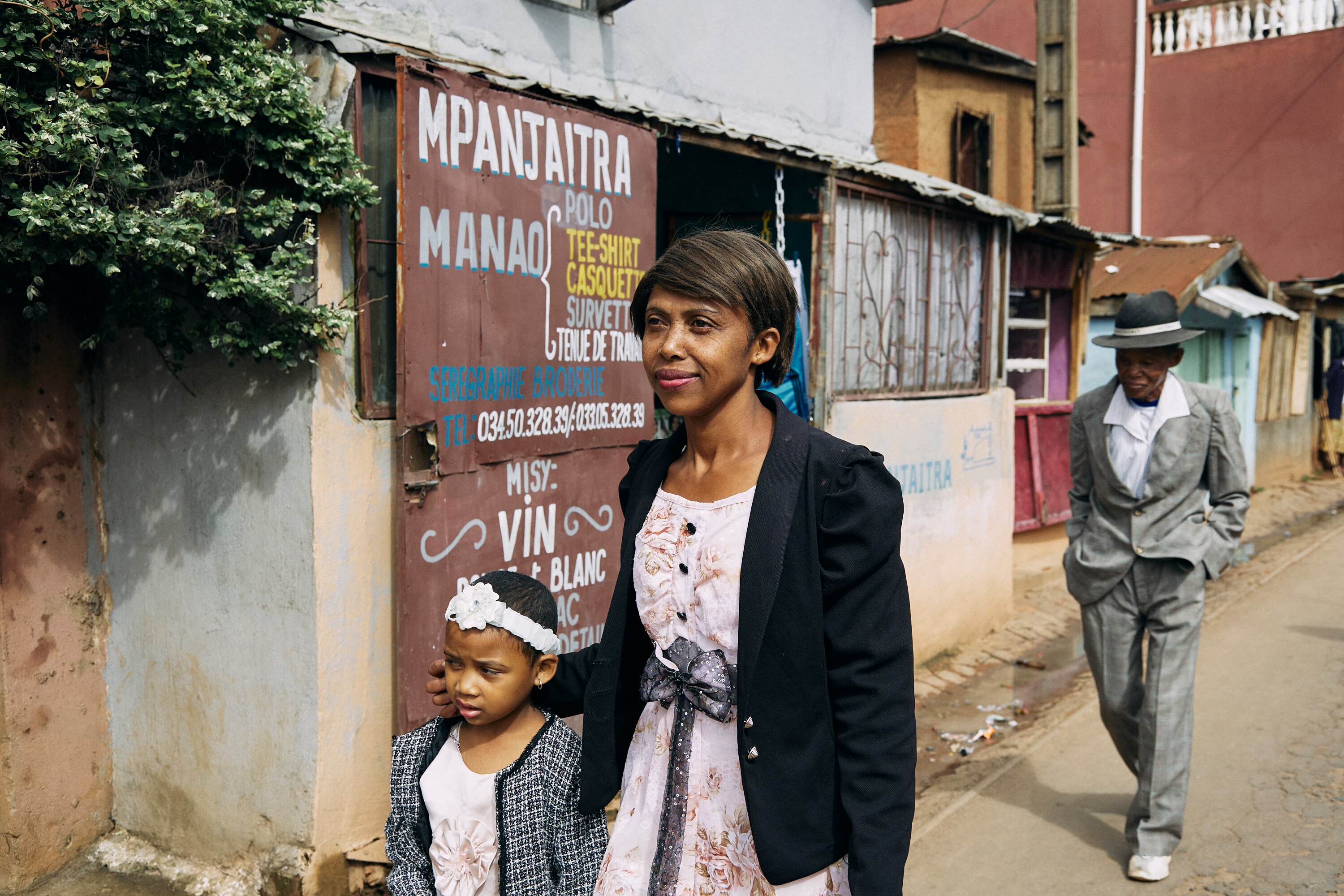 A woman and young girl walk along a street with an older man a few paces behind. They're all in their Sunday best. 