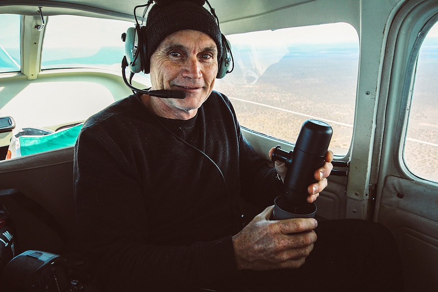 A photographer in black clothing sits on a light plane holding a camera.