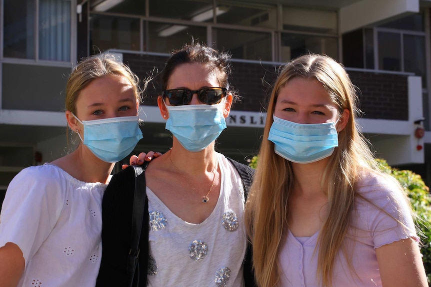 A mother stands posing for a photo outside flanked by her two teenage daughters, all wearing facemasks.