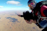 A young Indigenous man skydiving with a young woman. The man is smiling and gesturing to Uluru, pictured below.