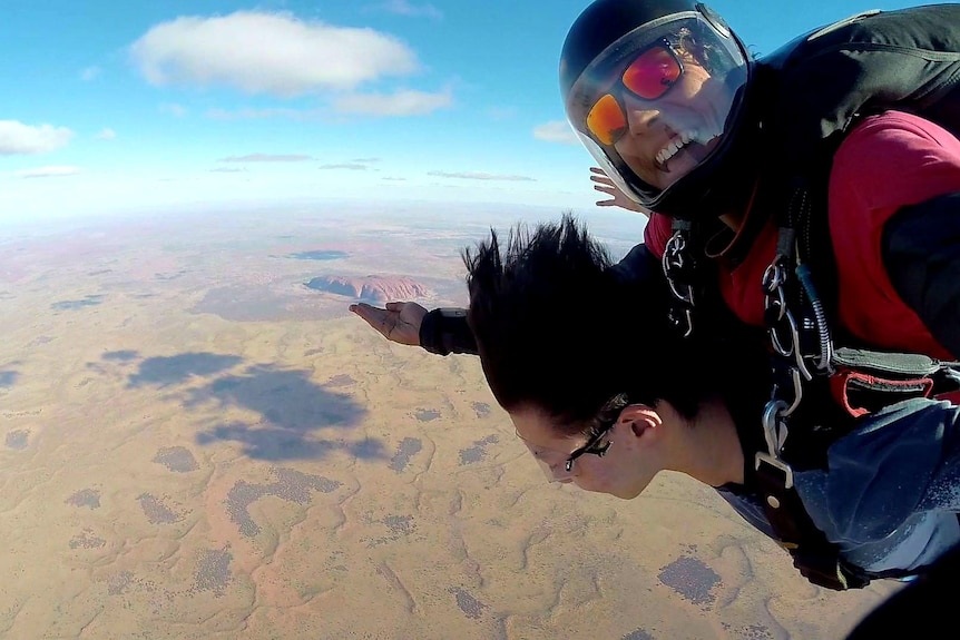 A young Indigenous man skydiving with a young woman. The man is smiling and gesturing to Uluru, pictured below.