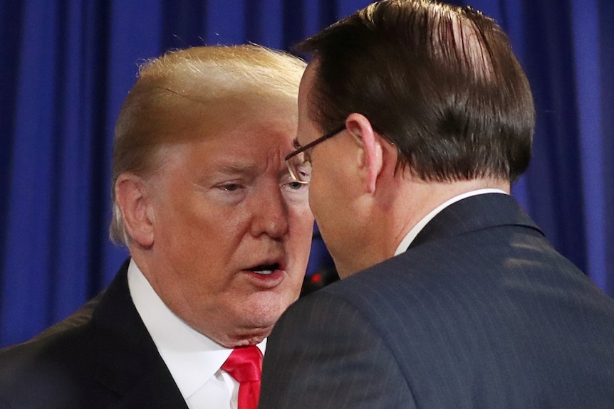 Two men lean in to move their heads closer to talk while standing in front of a blue drape