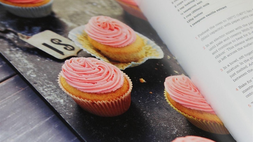 A cookbook opened to a page showing a photo of cupcakes with swirled pink icing.