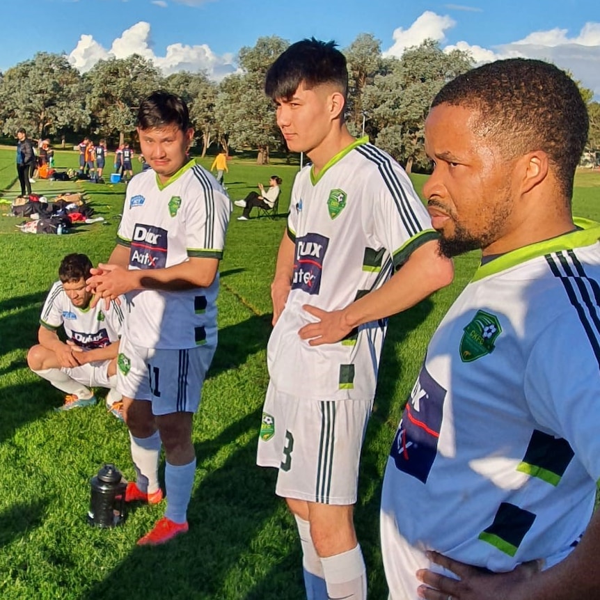 Men wearing football kits stand on the sideline of a football pitch.