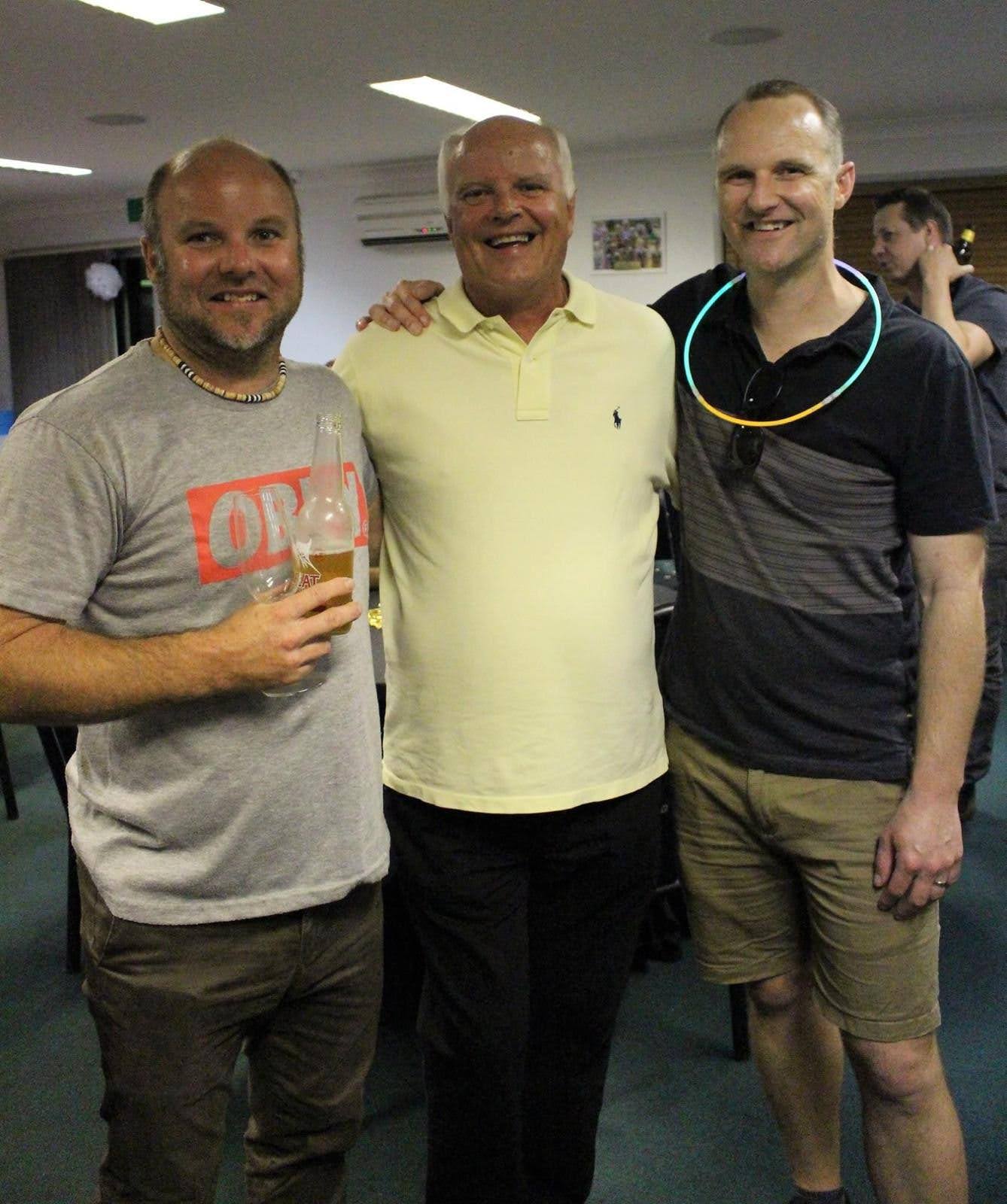 Three smiling men, one older, with their ams around each other at a venue.