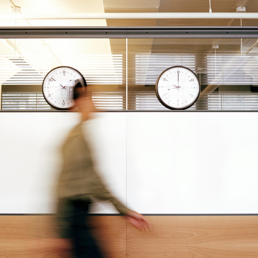 A blurry image of a human figure walking past a row of clocks in a corridor
