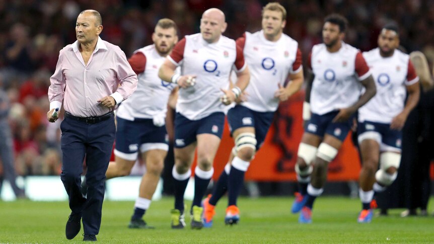 Eddie Jones runs in a shirt and trousers as his England team run a drill in the background