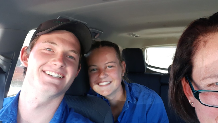 A teen boy and a teen girl, both dressed in blue shirts, smile from the front and back seat of a car.
