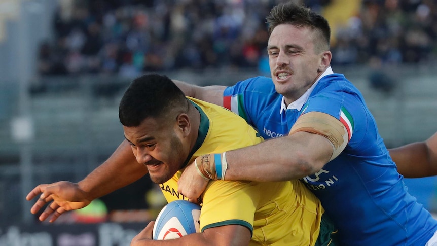 A man wearing blue tries to tackle a man carrying a rugby ball in yellow with his arms around his shoulders