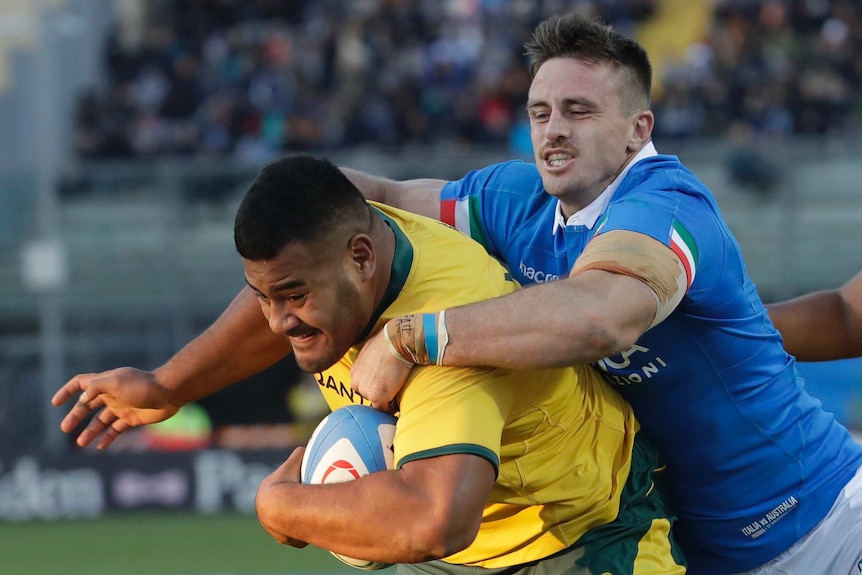A man wearing blue tries to tackle a man carrying a rugby ball in yellow with his arms around his shoulders