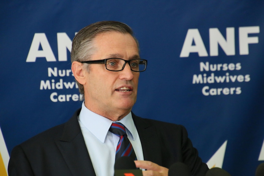 A headshot of Mark Olson wearing a suit and tie in front of a blue sign with white ANF writing.