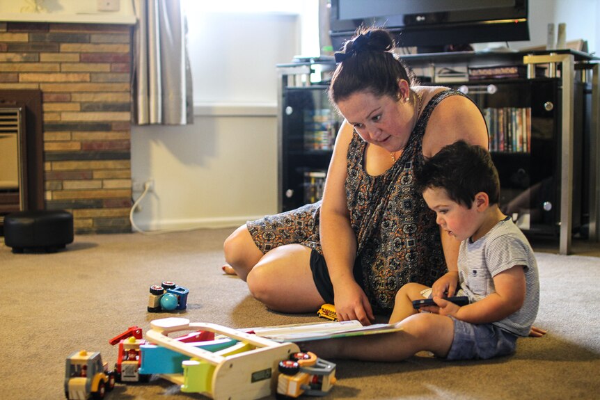Jess McGurk at home with her two-year-old son Tom sitting on the lounge room floor reading.
