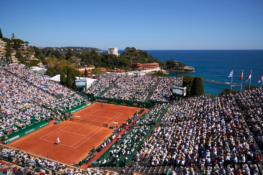 An overhead shot of a match at a tennis tournament in sunshine with packed crowds and the ocean in the background.