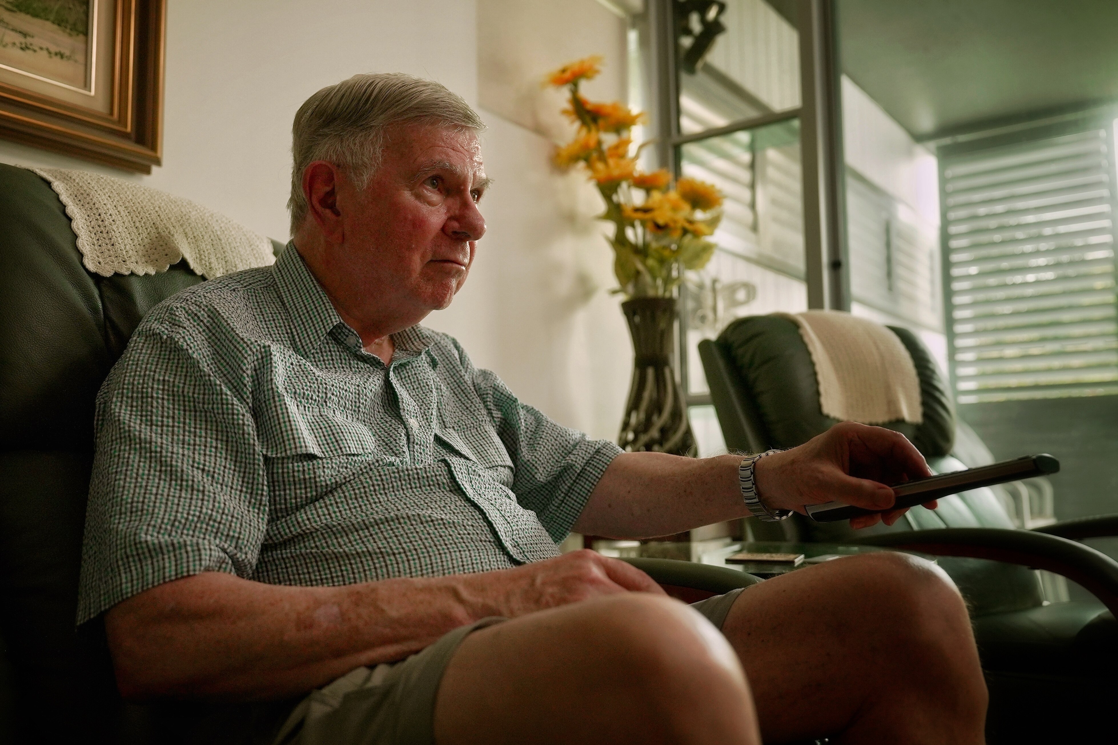 Un hombre blanco mayor con el pelo corto y gris está sentado en su sofá, con la camisa abotonada y sosteniendo el control remoto de un televisor.