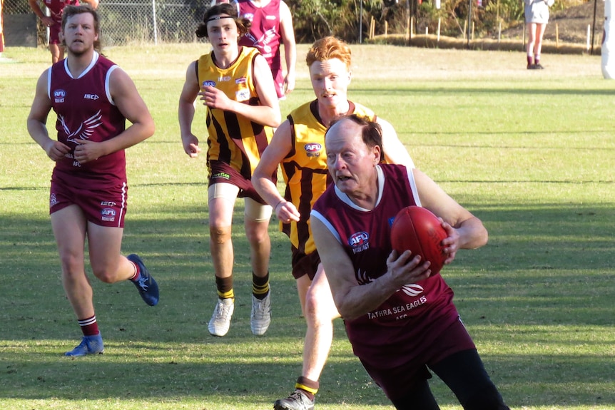 A senior man in maroon jumper and shorts runs away with an Aussie Rules football, with teammates and opponents behind