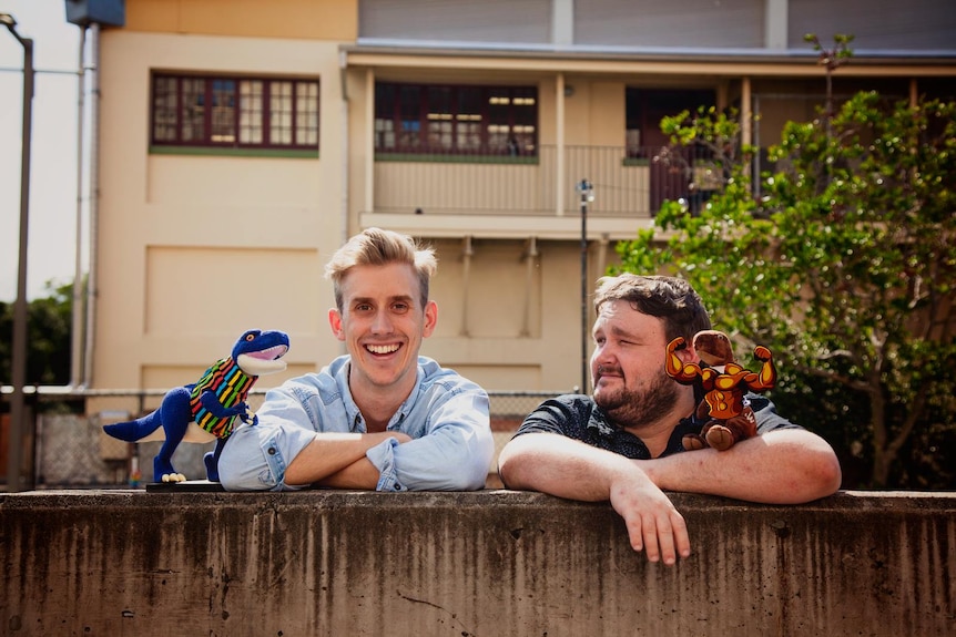 Two men lean on a wooden fence with stuffed toy dinosaurs beside them